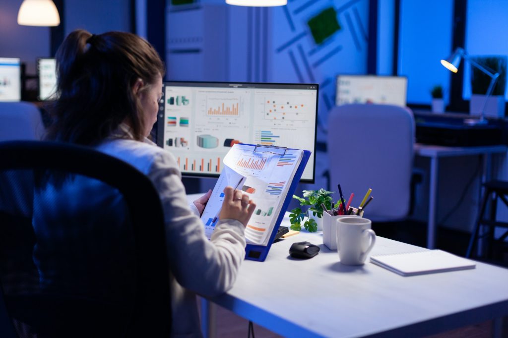 Freelancer woman comparing graphics from clipboard with grafics from computer